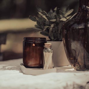 a brown glass jar, a white vase, and a small glass bottle on a table, with a plant in the background.