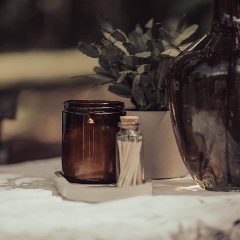 a brown glass jar, a white vase, and a small glass bottle on a table, with a plant in the background.