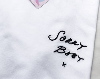 May include: White t-shirt with black embroidered text that says "Sorry Bro" with a small black cross underneath.