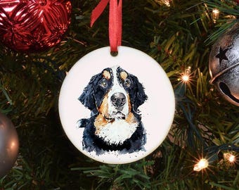 Può includere: Ornamento natalizio rotondo in ceramica con un ritratto ad acquerello di un bovaro del Bernese. Il cane ha il pelo nero, bianco e marrone. L'ornamento è appeso a un nastro rosso su un albero di Natale.
