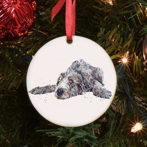 Könnte beinhalten: Ein runder weißer Weihnachtsbaumschmuck aus Keramik mit einem Aquarellbild eines liegenden grauen Hundes. Der Schmuck hat ein rotes Band, das oben befestigt ist.