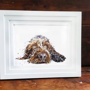 Puede incluir: Pintura a la acuarela de un perro en un marco blanco. El perro tiene pelaje marrón y negro, con una mirada enfocada. La obra de arte se exhibe sobre una superficie de madera, agregando un toque rústico al retrato de la mascota.