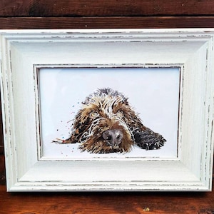 May include: A framed watercolor print of a dog with brown and black fur, lying down. The dog's eyes are open, and it has a dark nose. The frame is white with a distressed finish, set against a dark wood background.