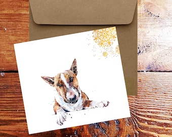 Puede incluir: Una tarjeta blanca con una ilustración de acuarela de un bull terrier marrón y blanco asomándose por el borde de la tarjeta. El perro tiene la nariz negra y los ojos marrones. El fondo de la tarjeta es blanco con copos de nieve dorados en la esquina superior derecha.