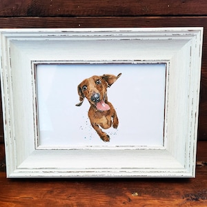 May include: A framed watercolor print of a dachshund dog with a surprised expression. The dog is brown with a pink tongue sticking out. The frame is white with a distressed finish, set against a dark wood background.