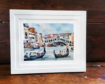 May include: Watercolor painting of Venice in a white frame. The artwork depicts gondolas on the water, buildings, and the Rialto Bridge. The painting has a blue sky and is framed in a white rectangular frame.