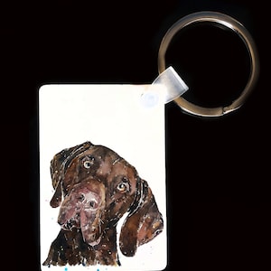 May include: A watercolour painting of a dog's head on a white rectangular keyring. The dog is brown and white and is looking to the side with its tongue slightly out. The keyring has a silver ring attached.
