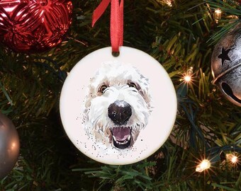 May include: A circular Christmas tree decoration with a watercolour image of a happy, fluffy white dog with a black nose and open mouth. The ornament is suspended by a red ribbon, set against a festive Christmas tree.