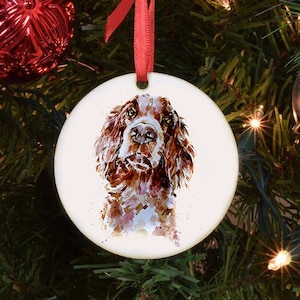 May include: A round white ceramic ornament with a watercolor painting of a brown and white dog. The dog is looking directly at the viewer with its tongue slightly sticking out. The ornament is hanging from a red ribbon.