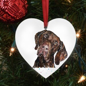 Puede incluir: Adorno navideño de cerámica blanca en forma de corazón con una pintura de acuarela de la cabeza de un perro marrón y negro. El perro está mirando hacia la izquierda de la imagen.