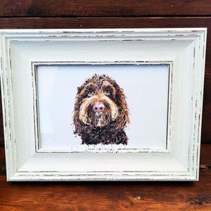 Op de afbeelding: Een ingelijste aquarelprint van een bruine en tan hond met een wit verweerde lijst. De vacht van de hond is gedetailleerd met penseelstreken en de lijst heeft een rustieke, verouderde uitstraling. Het kunstwerk wordt op een houten oppervlak getoond.