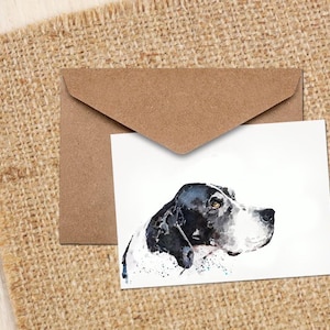 May include: A watercolour greeting card with a black and white dog portrait, alongside a brown envelope. The card is placed on a textured, tan hessian background, creating a rustic aesthetic.