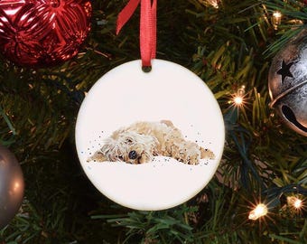 Pode incluir: Enfeite de Natal redondo e branco com uma ilustração em aquarela de um cão marrom claro deitado. O enfeite pende de uma fita vermelha contra um fundo de árvore de Natal decorada.