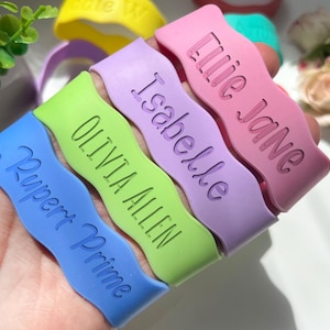 May include: A collection of wavy silicone wristbands in blue, lime green, lavender, and pink. Each band features debossed text including the names Rupert Prime, Olivia Allen, Isabelle, and Lilie Jane, with additional yellow and teal bands visible.