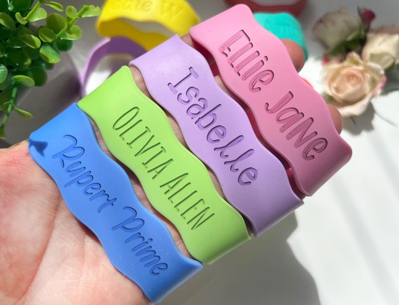 May include: A collection of wavy silicone wristbands in blue, lime green, lavender, and pink. Each band features debossed text including the names Rupert Prime, Olivia Allen, Isabelle, and Lilie Jane, with additional yellow and teal bands visible.