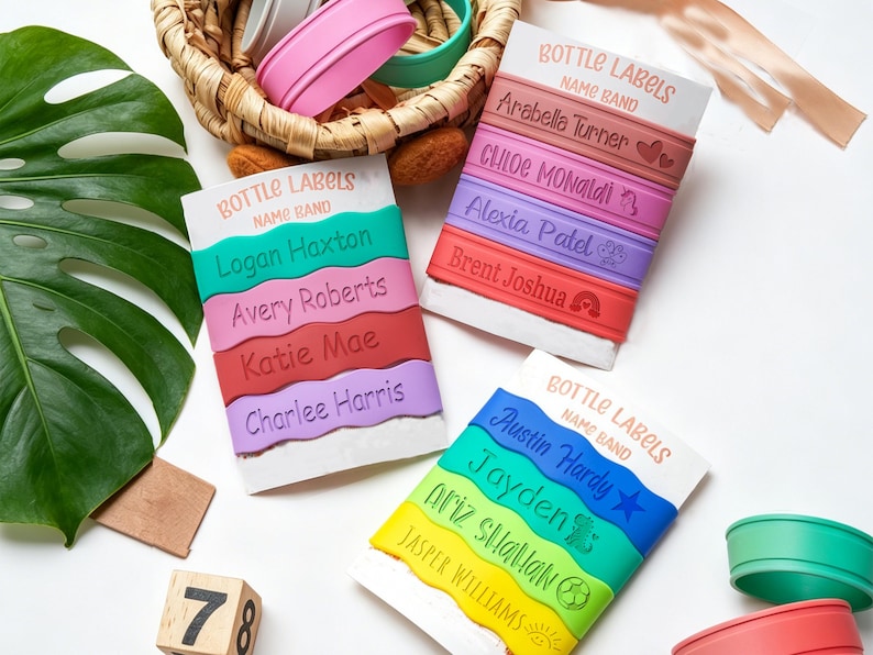 May include: Three white display cards labeled BOTTLE LABELS NAME BAND, each holding four colorful silicone bands. The bands feature engraved names like Logan Haxton, Arabella Turner, and Austin Hardy, alongside small icons like hearts, stars, and sports balls.