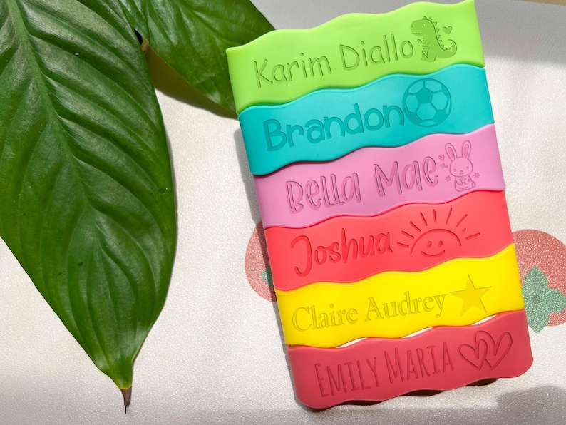May include: Six colorful silicone name tags with wavy edges. The names are "Karim Diallo", "Brandon", "Bella Mae", "Joshua", "Claire Audrey", and "Emily Maria". Each tag has a different graphic: a dinosaur, a soccer ball, a bunny, a sun, a star, and a heart.