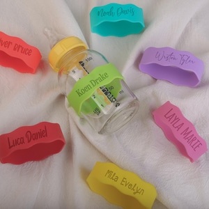 May include: A set of ten colorful silicone bottle labels with names printed on them. The labels are in various colors, including red, pink, purple, yellow, and green. The labels are designed to be used on baby bottles.