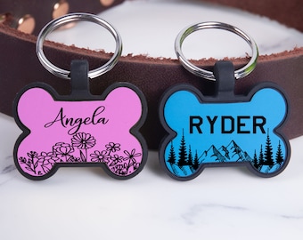 Puede incluir: Dos etiquetas personalizadas con forma de hueso para perro. La etiqueta rosa tiene el nombre "Angela" y un diseño floral. La etiqueta azul tiene el nombre "RYDER" y una escena de montaña.