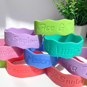 May include: A collection of colorful silicone wristbands with wavy edges, each featuring embossed names and icons. The bracelets appear in shades of lime green, lavender, pink, teal, blue, and coral, with text including Saylor, Ace G., Mr. Amir, Mason, and Cara Ker.