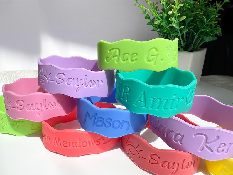 May include: A collection of colorful silicone wristbands with wavy edges, each featuring embossed names and icons. The bracelets appear in shades of lime green, lavender, pink, teal, blue, and coral, with text including Saylor, Ace G., Mr. Amir, Mason, and Cara Ker.