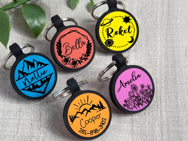 May include: A collection of five colorful pet ID tags with black rubber surrounds and silver key rings. Each tag features a different design, including names like "Nellie," "Bella," "Roket," "Amelia," and "Cooper," along with various graphics and a phone number.