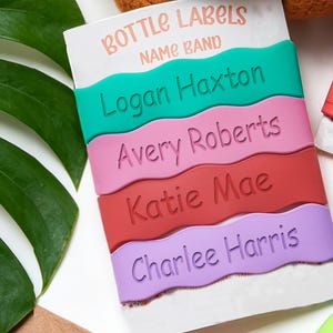 May include: A set of four wavy silicone bottle labels in teal, pink, red, and purple, each featuring a debossed name: Logan Haxton, Avery Roberts, Katie Mae, and Charlee Harris. The labels are displayed on a white card labeled BOTTLE LABELS NAME BAND.
