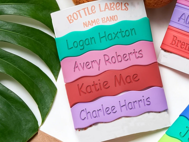 May include: A set of four wavy silicone bottle labels in teal, pink, red, and purple, each featuring a debossed name: Logan Haxton, Avery Roberts, Katie Mae, and Charlee Harris. The labels are displayed on a white card labeled BOTTLE LABELS NAME BAND.