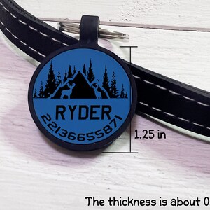 May include: A blue and black pet tag with the name "RYDER" and a phone number. The tag is round, 1.25 inches in diameter, and features a mountain and forest design. The tag is attached to a black leather collar.