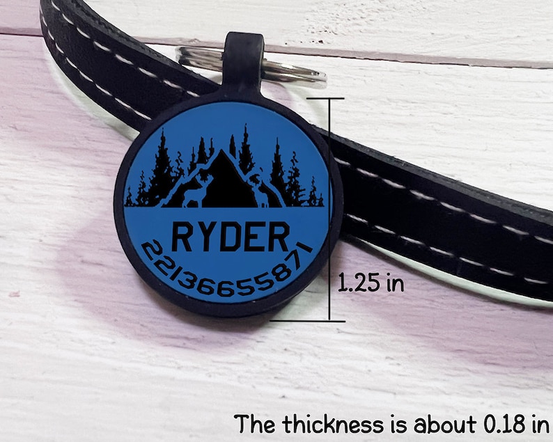 May include: A blue and black pet tag with the name "RYDER" and a phone number. The tag is round, 1.25 inches in diameter, and features a mountain and forest design. The tag is attached to a black leather collar.