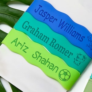 May include: Three wavy silicone bands in blue, teal, and lime green. Each band features a custom name engraved in dark blue or green: Jasper Williams with a dolphin, Graham Romer with a cow, and Ariz Shahan with a soccer ball graphic.