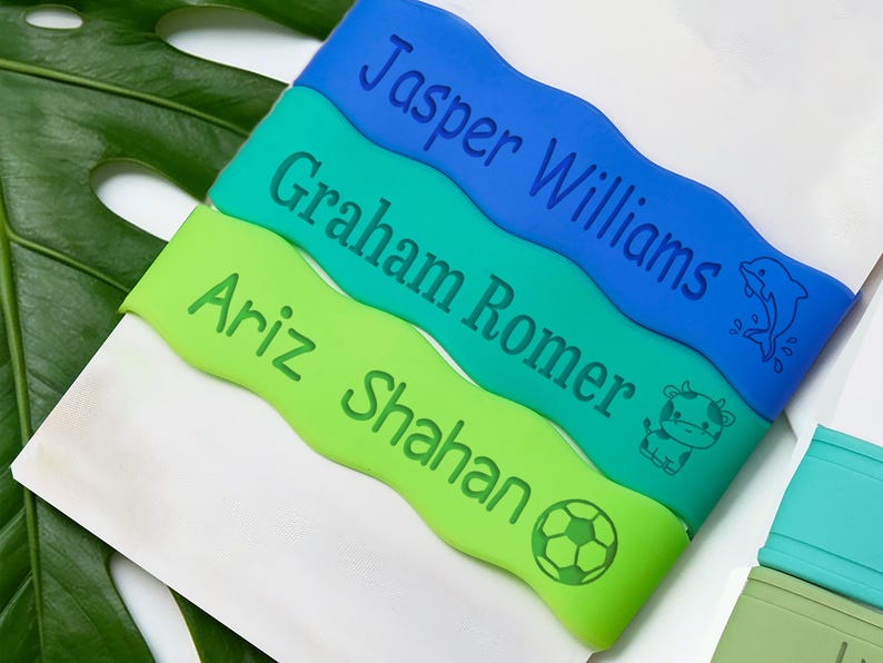 May include: Three wavy silicone bands in blue, teal, and lime green. Each band features a custom name engraved in dark blue or green: Jasper Williams with a dolphin, Graham Romer with a cow, and Ariz Shahan with a soccer ball graphic.