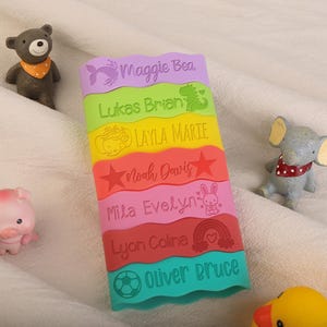 May include: A rectangular, multi-colored silicone strip with wavy edges featuring six horizontal sections in purple, lime green, yellow, orange, pink, red, and teal, each engraved with a name and icon: Maggie Bea, Lukas Brian, Layla Marie, Noah Davis, Mila Evelyn, Lyon Coline, and Oliver Bruce.