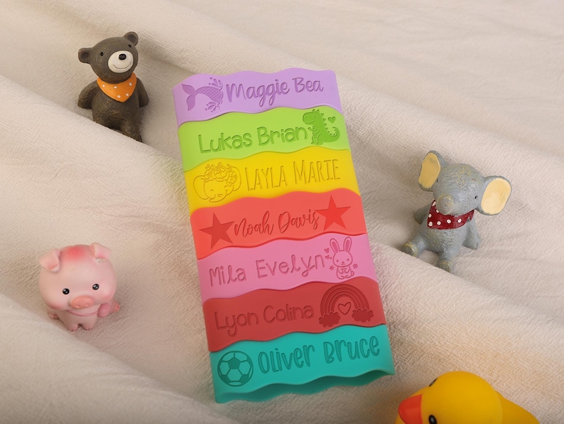 May include: A rectangular, multi-colored silicone strip with wavy edges featuring six horizontal sections in purple, lime green, yellow, orange, pink, red, and teal, each engraved with a name and icon: Maggie Bea, Lukas Brian, Layla Marie, Noah Davis, Mila Evelyn, Lyon Coline, and Oliver Bruce.