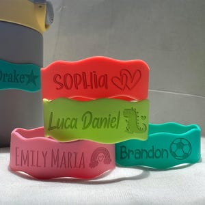 May include: Five colorful silicone wristbands with personalized names and designs. The wristbands are stacked on top of each other. The names on the wristbands are "Koen Drake", "Sophia", "Luca Daniel", "Emily Maria", and "Brandon".