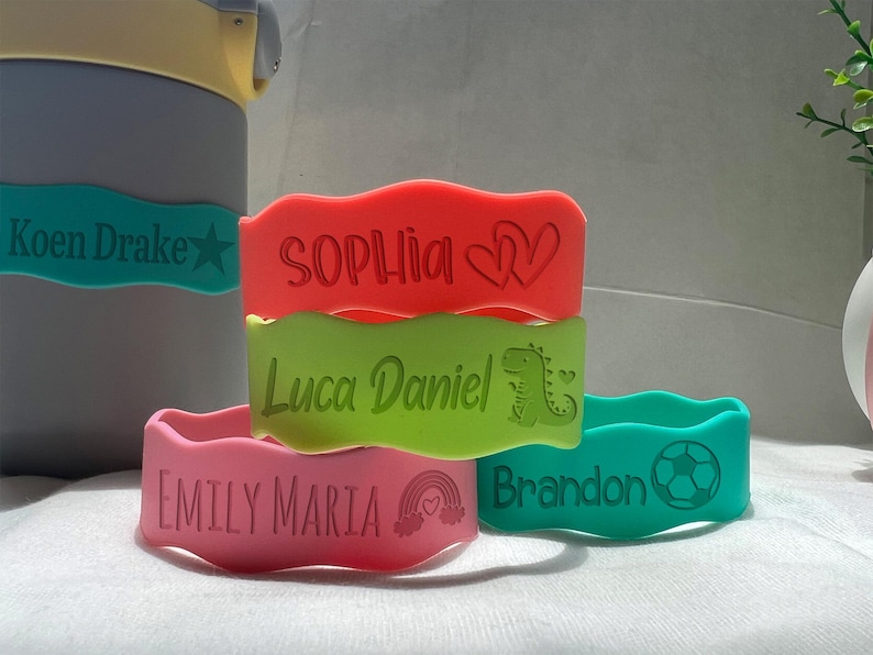 May include: Five colorful silicone wristbands with personalized names and designs. The wristbands are stacked on top of each other. The names on the wristbands are "Koen Drake", "Sophia", "Luca Daniel", "Emily Maria", and "Brandon".