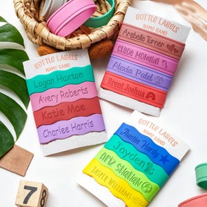 Może przedstawiać: Trzy białe karty ekspozycyjne z napisem BOTTLE LABELS NAME BAND, każda z czterema kolorowymi silikonowymi opaskami. Opaski mają wygrawerowane imiona, takie jak Logan Haxton, Arabella Turner i Austin Hardy, oraz małe ikony, takie jak serca i gwiazdki.