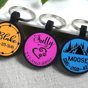 May include: Three personalized pet ID tags with black rubber surrounds and silver key rings. The tags feature names and phone numbers on orange, pink, and blue backgrounds. One tag has stars, another a heart and paw prints, and the third a mountain scene.