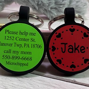 May include: Two round pet ID tags. One is green with black text: "Please help me" and contact information. The other is red with the name "Jake" and heart-shaped cutouts. Both have black loops and silver key rings.