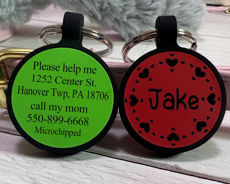 May include: Two round pet ID tags. One is green with black text: "Please help me" and contact information. The other is red with the name "Jake" and heart-shaped cutouts. Both have black loops and silver key rings.