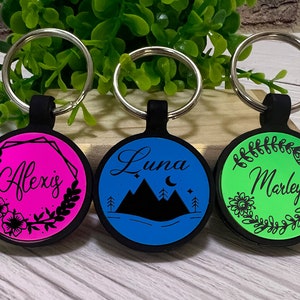 May include: Three personalized keychains with black rubber holders and silver key rings. The keychains have round, colorful faces with names and designs: Alexis on pink, Luna with mountains on blue, and Marley on green.