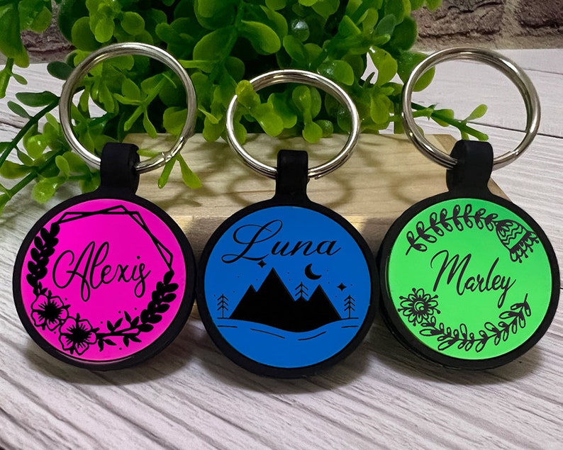 May include: Three personalized keychains with black rubber holders and silver key rings. The keychains have round, colorful faces with names and designs: Alexis on pink, Luna with mountains on blue, and Marley on green.