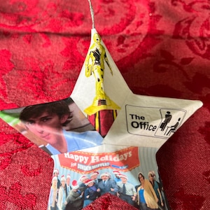 May include: A white star-shaped ornament with a collage of images from the TV show "The Office". The ornament features a picture of Jim Halpert, the "Dunder Mifflin" logo, and the text "Happy Holidays".