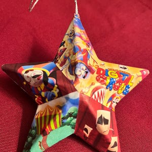 May include: A colorful star-shaped ornament made from paper with a silver string for hanging. The ornament features images of clowns, a circus tent, and the text "The Amazing Digital Circus".