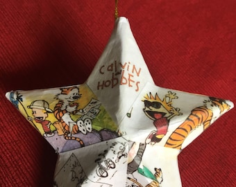 May include: A white star-shaped ornament with a red, orange, and black cartoon image of Calvin and Hobbes. The ornament has a gold string for hanging and the words "Calvin and Hobbes" are printed on the top.
