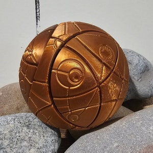 Puede incluir: Una esfera dorada metálica con un patrón geométrico, descansando sobre tres piedras grises.