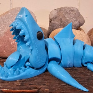 Può includere: Un modello di squalo articolato stampato in 3D di colore blu con la bocca aperta e denti affilati. Il modello è posizionato su una superficie di legno con rocce sullo sfondo.