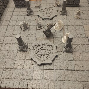 K&ouml;nnte beinhalten: Ein graues Tabletop-Spielset mit Miniaturfiguren und einer steinartigen Textur. Das Set enth&auml;lt eine runde Basis mit einem Pentagramm-Design, S&auml;ulen und Figuren verschiedener Charaktere.