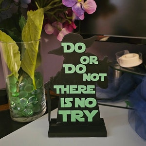 以下が含まれることがあります： 緑色の文字で「Do or do not, there is no try」と書かれた架空のキャラクターの黒いシルエットです。