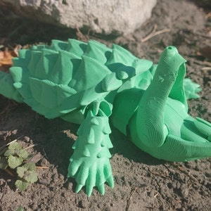 以下が含まれることがあります： 鮮やかな緑色の3Dプリントされたカメの置物。カメは、とげのある突起のある、詳細で質感のある甲羅を持っています。足と頭も複雑に設計されており、ユニークで芸術的な外観になっています。置物は土と小さな植物の上に置かれています。